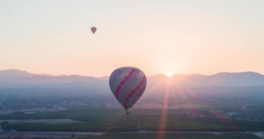 Several hot air balloons drift over a valley at sunrise with mountains in the background