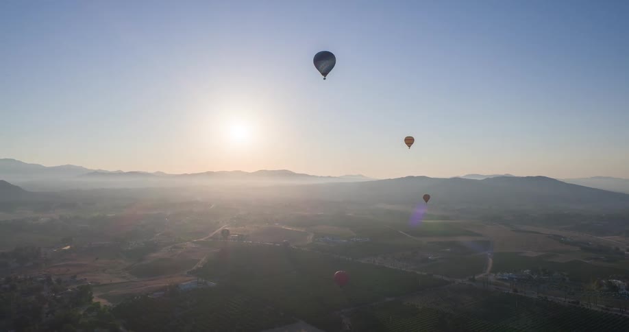 A view from a high angle shows a landscape with rolling hills and a hazy sunrise as three hot air balloons float in the clear sky above the ground