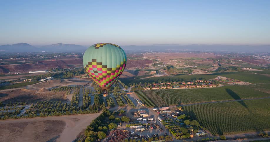 The video shows an aerial shot of a colorful hot air balloon with a teal pink and yellow checkered pattern it is floating over a rural landscape with vineyards and small towns in the distance