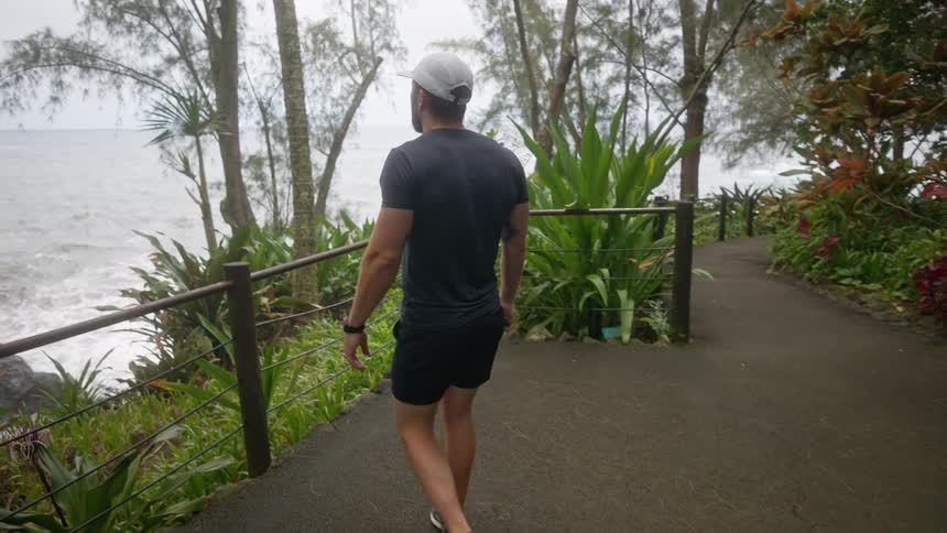 The video shows a man walking on a paved path surrounded by lush green foliage and trees he is wearing a dark shirt shorts and a baseball cap the man pauses to take a picture of the ocean with his phone which is visible on the left side of the frame