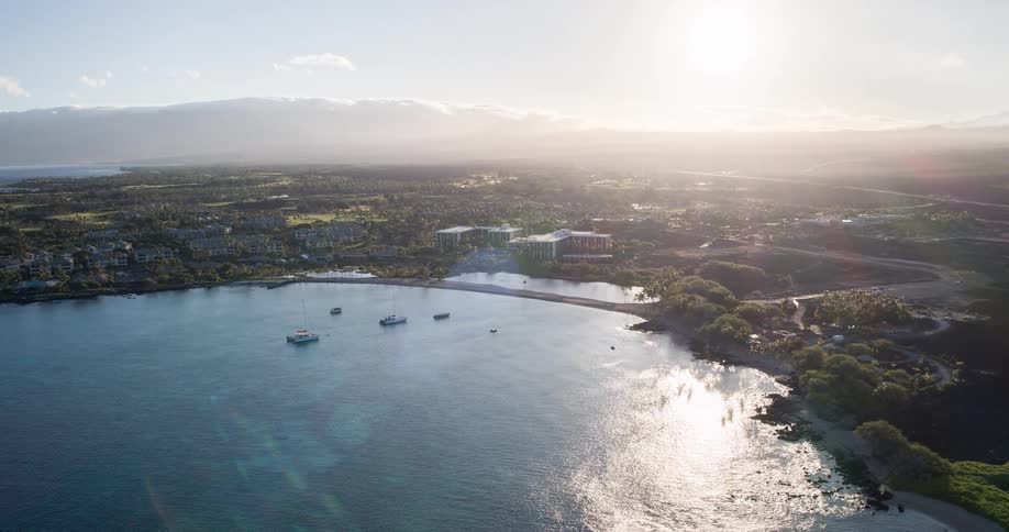 The video shows a wide aerial view of a coastline with a bay and a town the sun is shining brightly creating a haze over the landscape