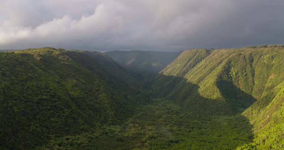 The video shows a wide aerial view of a lush green valley the camera pans across the valley showing the a cloudy sky and the green hills