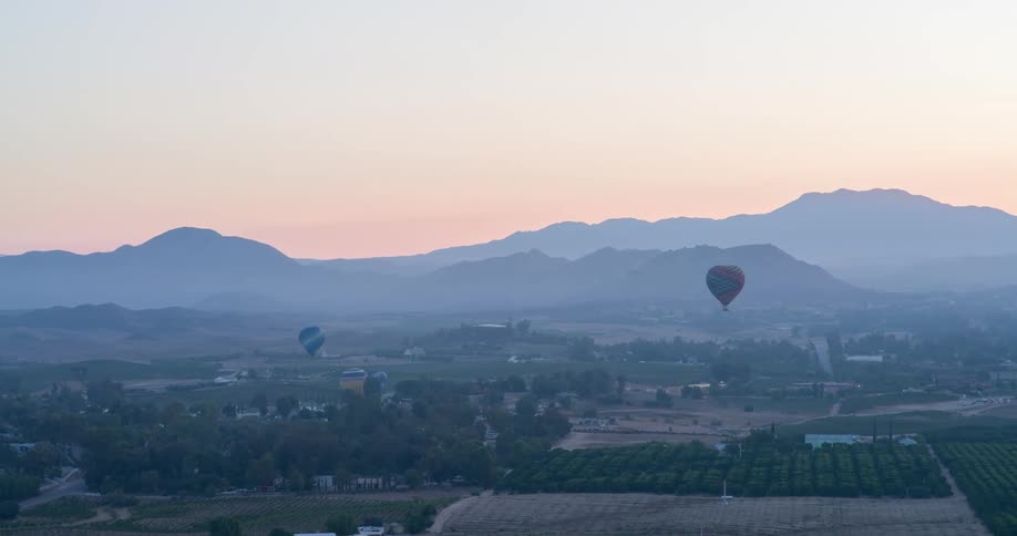 The video shows a scenic aerial view of a landscape with hills and valleys two hot air balloons are seen floating in the air against a hazy pinktinged sky
