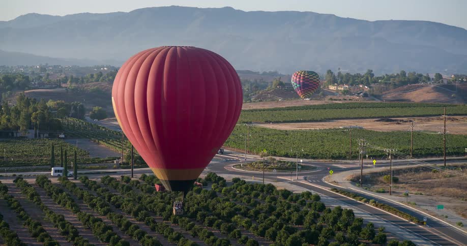 An aerial drone shot shows a red and orange hot air balloon floating over a landscape of vineyards and hills another multicolored hot air balloon is also visible in the distance