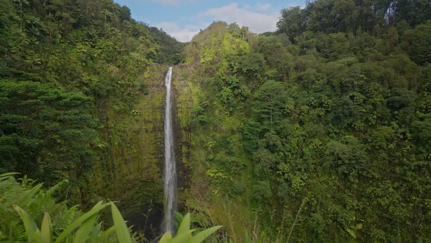 A beautiful tall waterfall cascading down a cliff face covered in lush green moss and foliage with the sky and clouds visible at the top