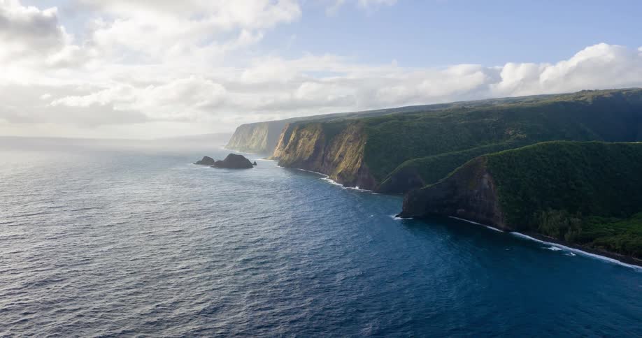 Sunlight illuminates a stunning rugged coastline stretching into the vast blue ocean under a cloudy sky