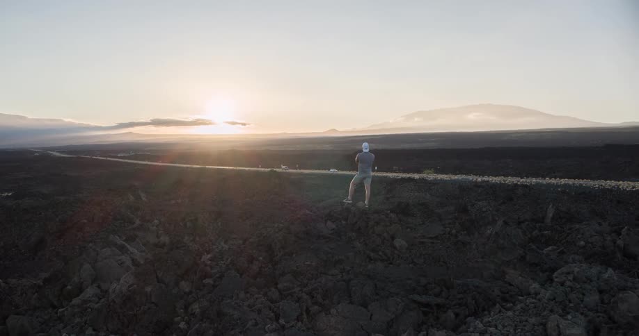A man wearing a white hat sneakers shorts and an ash tshirt stands on the peak of a rock in a rocky terrain staring at the setting sun