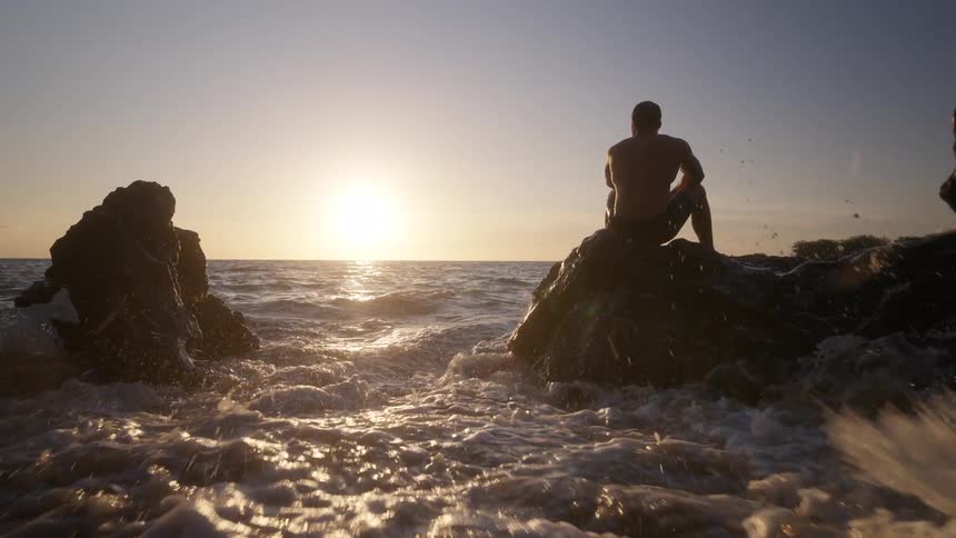 Man with his back turned sitting on a rock in the sea with waves coming towards him