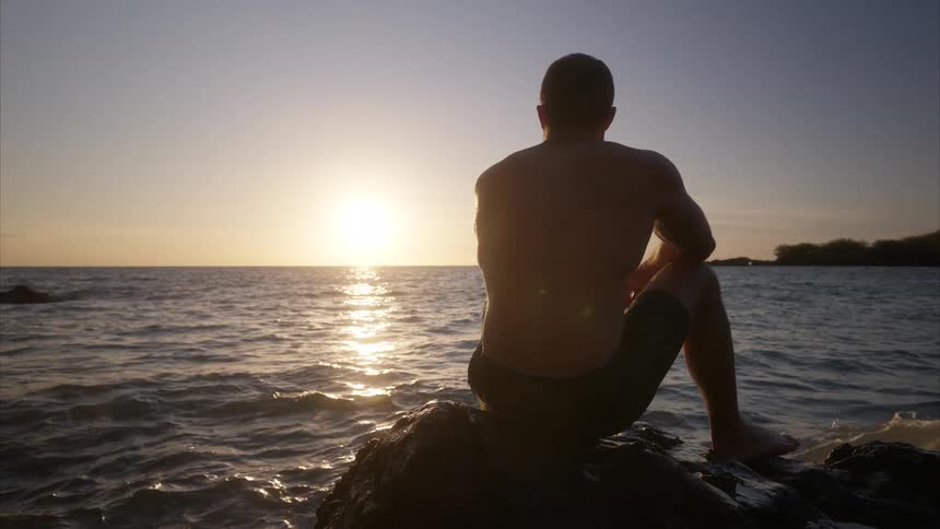 Man sitting on rocks along the coast watching waves roll into shore as the sun sets on the horizon