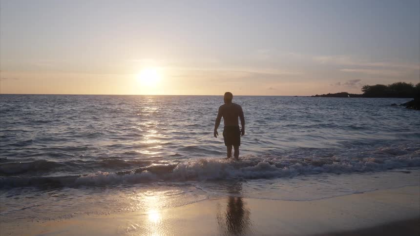 A man stands in the shallow waves of the ocean watching a serene sunset over the horizon