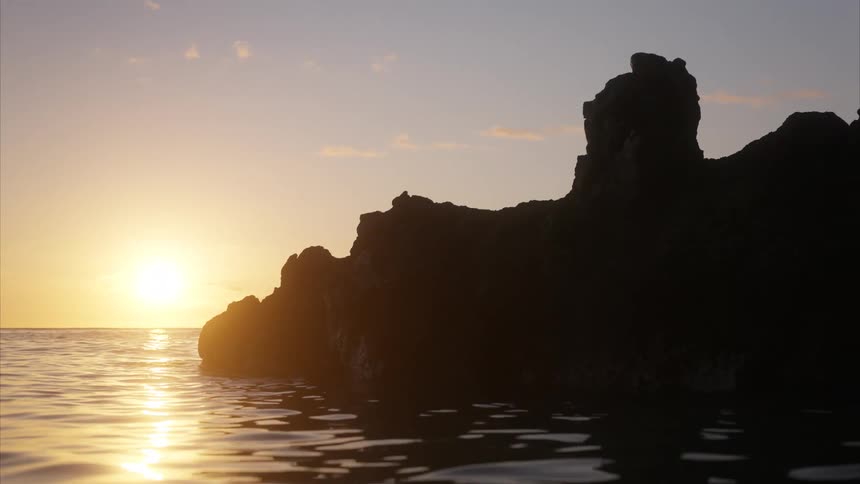 A large rock in the ocean water with the orange setting sun in the distance as the water is filled with ripples caused by waves