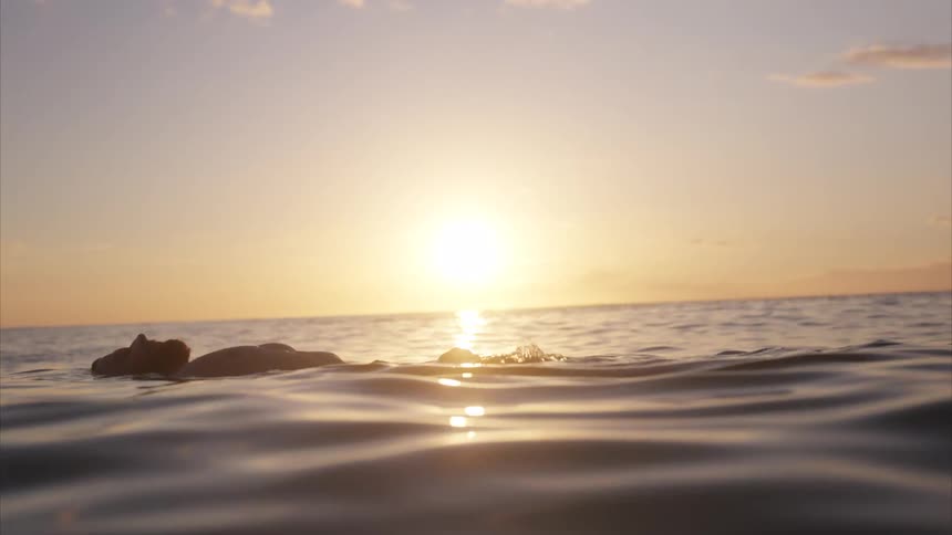 A person floats peacefully on their back in the calm ocean as the sun sets casting a golden glow over the water