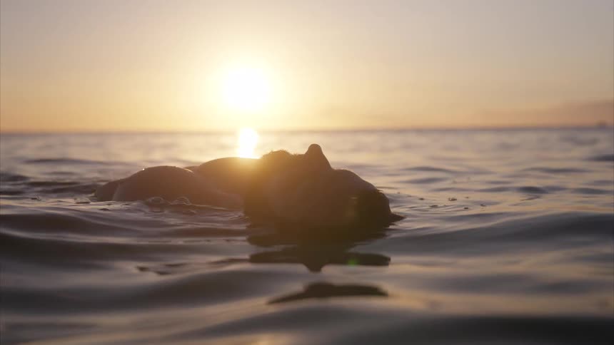 A person is floating on their back in the ocean at sunset with the suns reflection visible on the water