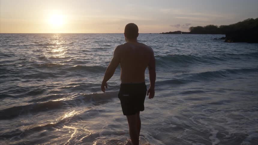 A man wearing trunks slowly walks into the ocean from the beach at sunset