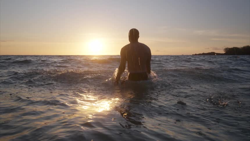 A shirtless man stands waistdeep in the ocean splashing the water with both hands as the waves create ripples with the orange setting sun in the distance