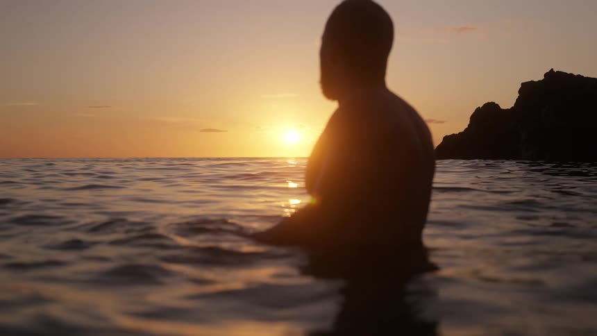 A shirtless man swims in the ocean near the coast submerged from the waist down with the orange setting sun in the distance