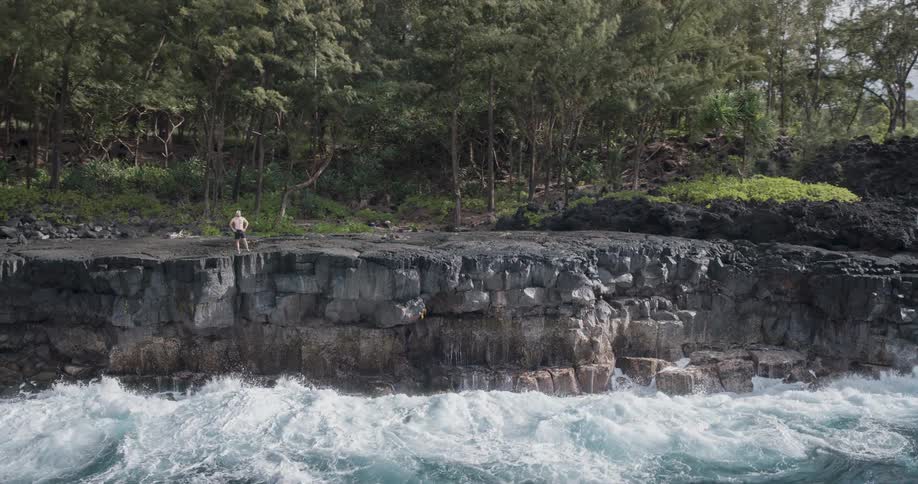 The man is watching the waves crash against the cliff