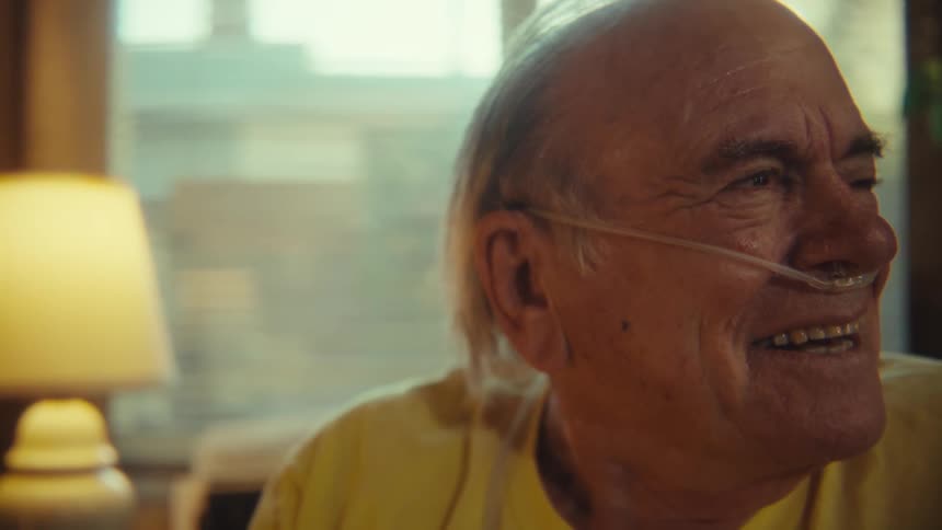 An elderly man sits on a chair in his living room wearing a nasal cannula laughing and excited at midday
