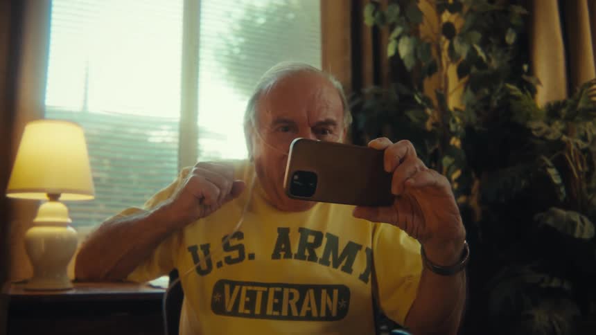 An elderly man is watching a video on the smartphone while sitting in a wheelchair near a window
