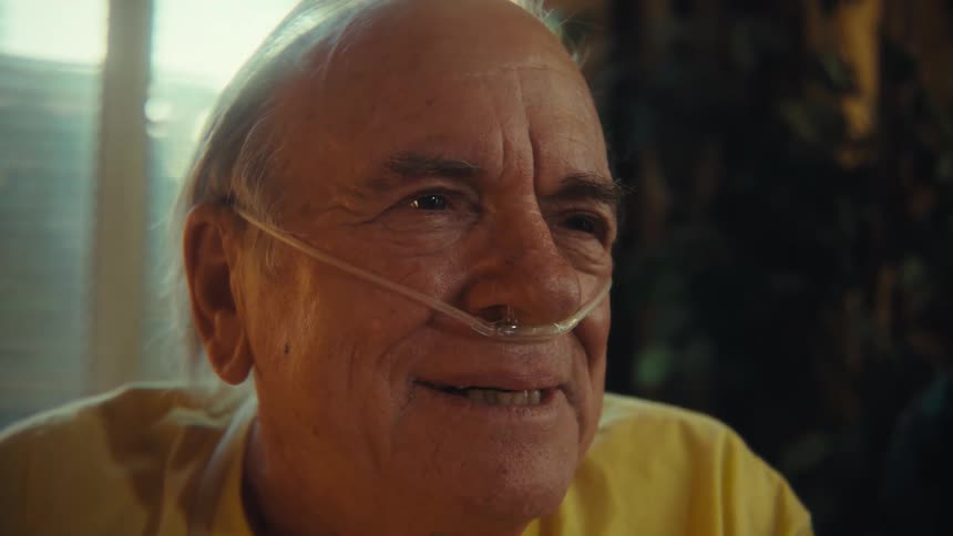 A man with white hair wearing a yellow shirt and a nasal cannula is looking to his left and smiling while a window and some plants are visible in the blurry background