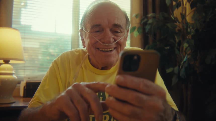 An elderly man in a wheelchair laughs as he looks at a smartphone in his hand