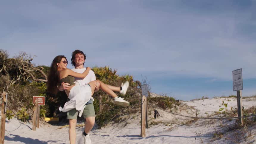 A smiling couple is on a sandy path with a man wearing a white tshirt and green shorts carrying a woman in his arms who is wearing a white dress over a dark top with the ocean and a cloudy sky in the background