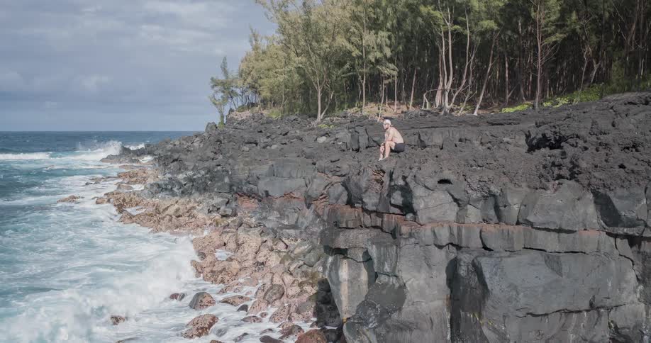 Man sitting on rocky cliff watching waves crash on shoreline