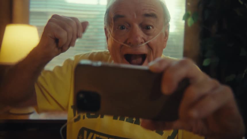 A joyful man wearing a yellow us army veteran tshirt and an oxygen tube is sitting in a chair looking at his smartphone waving and smiling joyfully