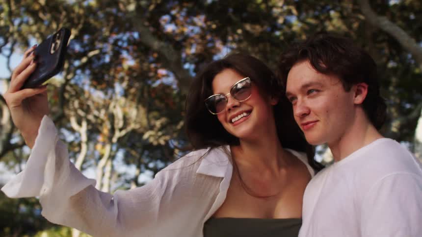 A young couple stops running in front of a twisted tree embrace while smiling and pose for a selfie on her cellphone