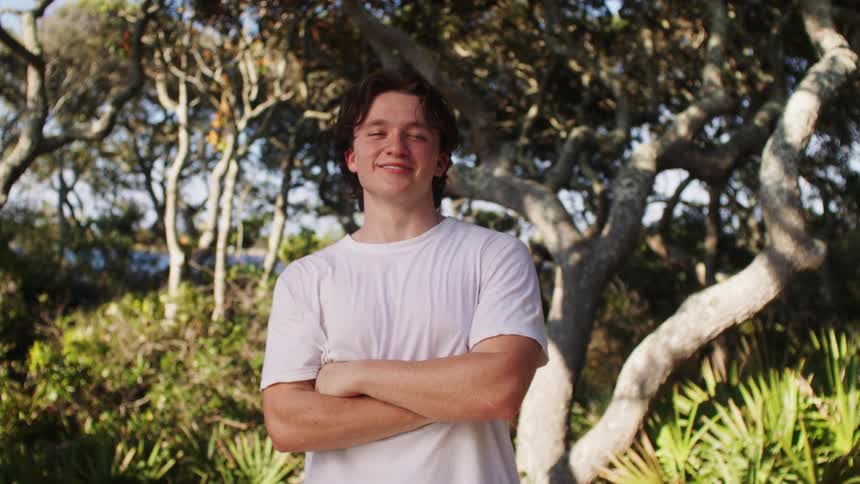 A young man with brown hair and a happy expression is standing with his arms crossed wearing a white tshirt and smiling at the viewer while standing in a forest with sun shining on the trees behind him