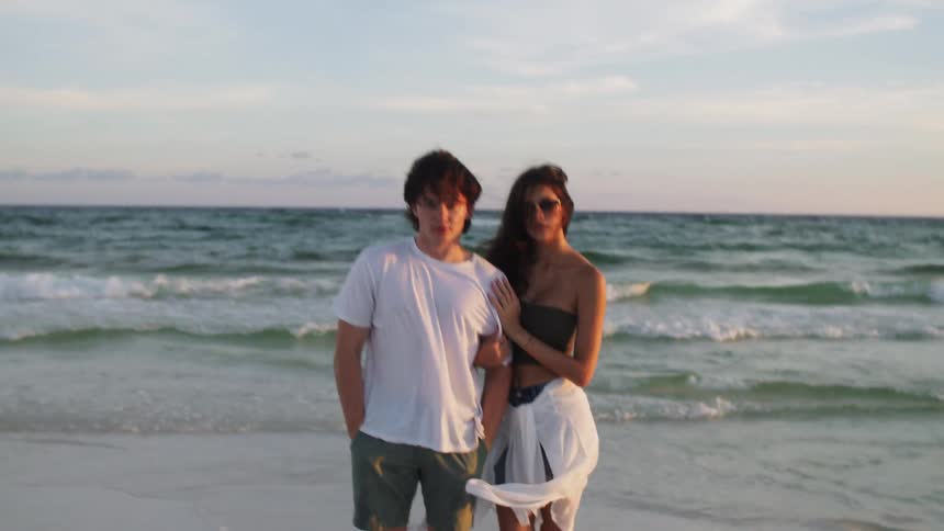 Young guy and young girls stand on a beach with their back facing the waves the guy is wearing a white tshirt and green shorts with his hands in his pockets the girl has long hair sunglasses tub top and a white wrap around her waist