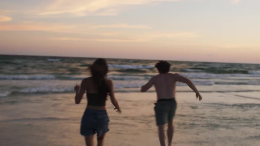 Young couple running on sand towards the ocean tossing shirts aside at sunset
