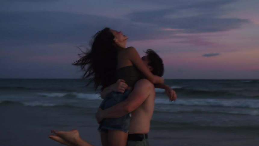 A couple is embracing on a beach with a man carrying a woman and spinning her around as the sun sets behind them
