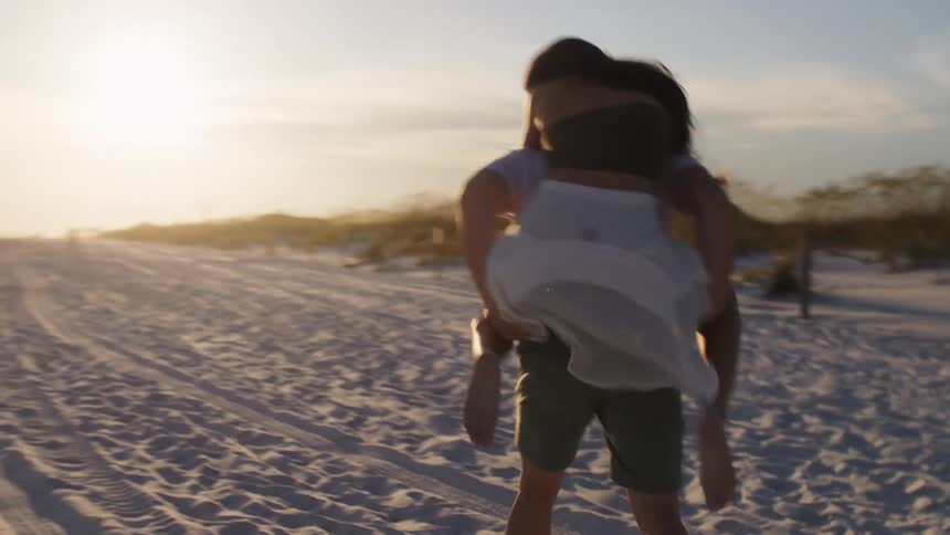 A couple having fun on the beach at sunset with the woman running behind the man then hopping on his back as he carries her while running on the sand both are laughing and enjoying themselves