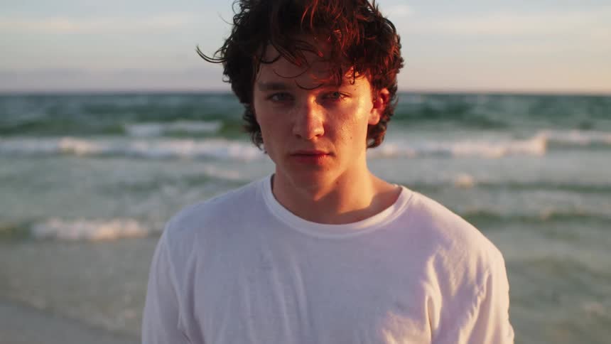 Youn man stands on the beach as wind blows his shirt and waves roll into shore