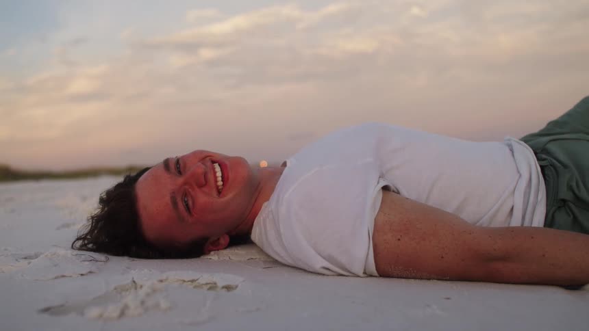 A young man lying on the beach sand during a vacation laughing at sunset