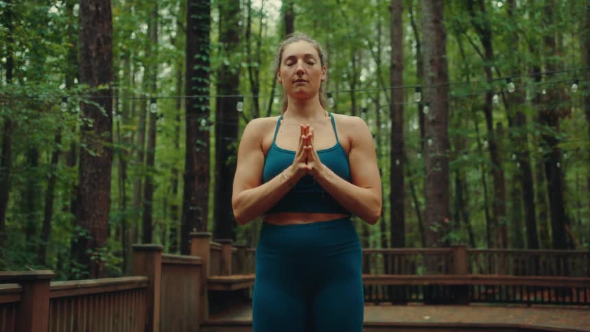 A young woman wearing blue tights and a blue top does yoga beside a cabin in the woods at midday