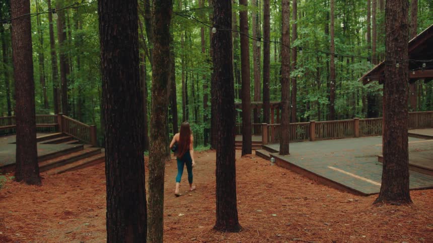 A woman with long brown hair wearing a blue workout outfit is walking across a pine needlecovered ground in a forest carrying a black yoga mat toward a wooden deck with string lights hanging overhead