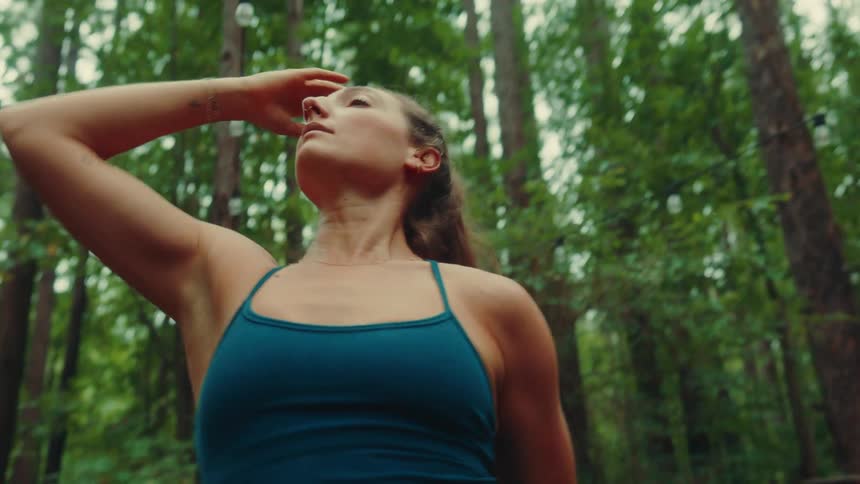A female yogi wearing a dark blue sports bra is stretching her arms and body in a side bend with her hair in a ponytail and a line of string lights hanging in the green forest behind her