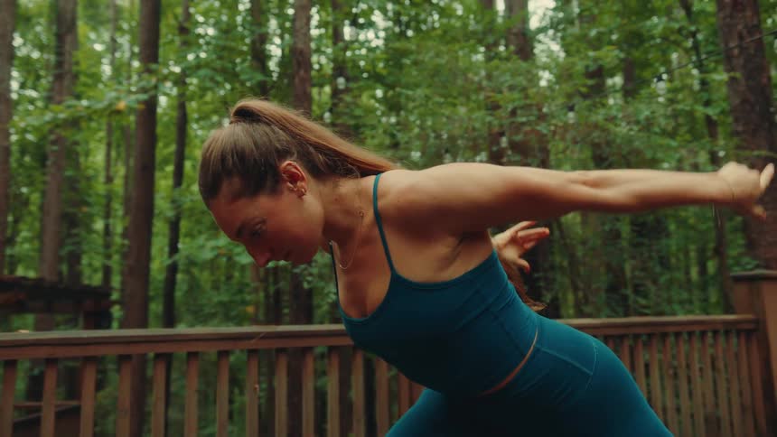 A female yogi with her hair in a ponytail is practicing yoga on a wooden deck in a dense forest doing a sun salutation with her arms stretched to the sky and then slowly moving her body to the side
