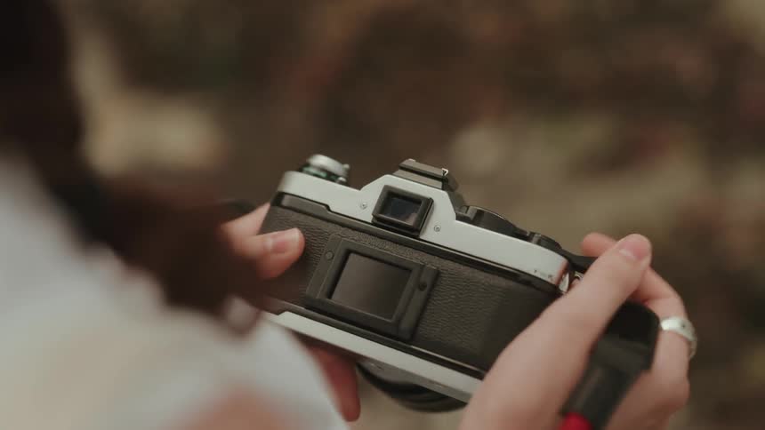 A closeup of a persons hands with two rings shows them holding a vintage silver and black 35 mm film camera against a blurry earthytoned background