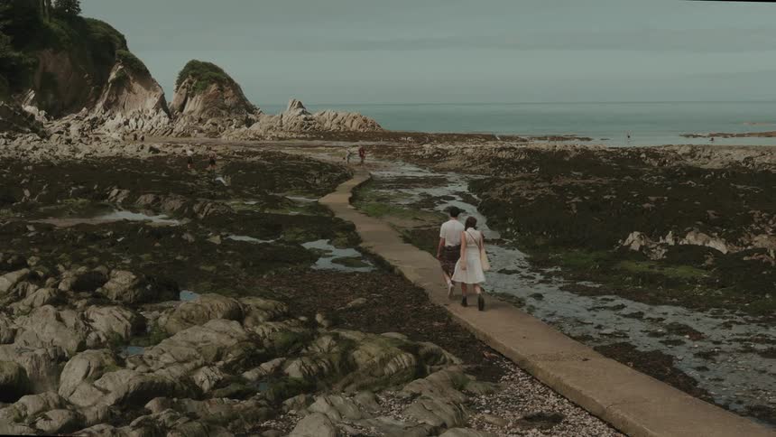 A man and woman walk along a narrow path through rocky tidal flats toward the sea