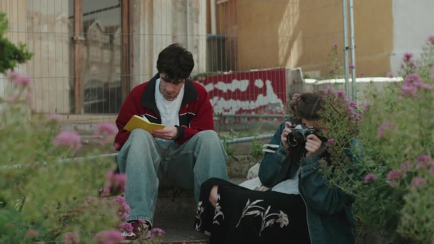 A young couple is sitting on outdoor steps surrounded by pink wildflowers with a man in a red and black jacket writing in a yellow notebook while a woman in a denim jacket takes a picture with a vintage 35mm film camera