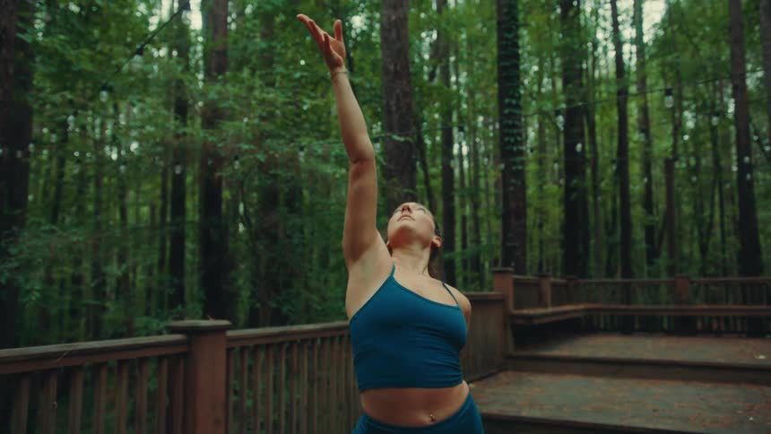 A female yogi wearing a dark blue sports bra and leggings is practicing yoga on a wooden deck in a forest transitioning from a lunge to a side bend with her arms stretching out and up