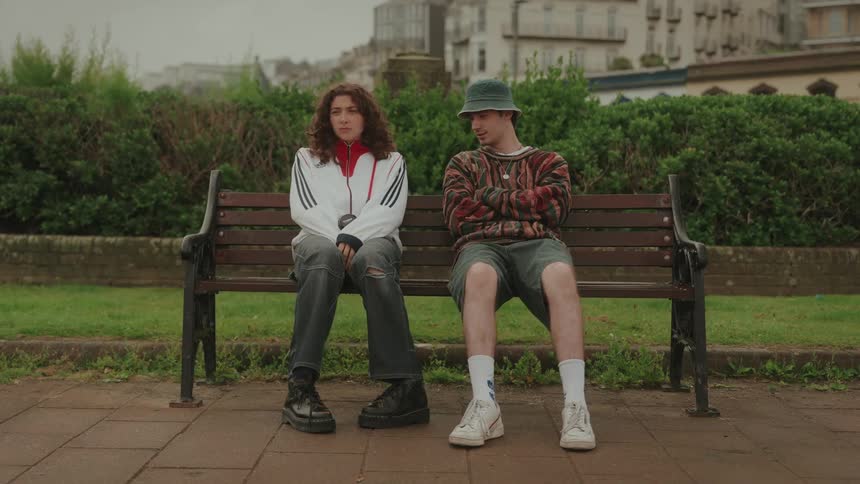Two people sit on a park bench looking in different directions with serious expressions