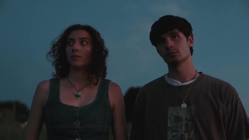 A man and woman with serious expressions are standing next to each other with the woman wearing a denim top and necklace and the man wearing a tshirt and necklace against a hazy deep blue sky