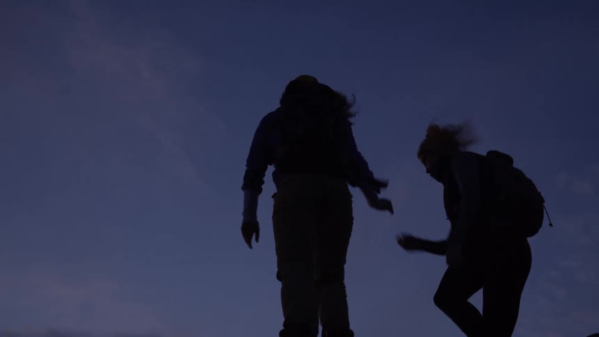 Two women hiking on a rocky terrain at sundown with one holding the other to help her climb up a rock both carrying backpacks