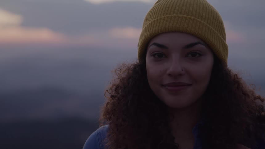 A young woman wearing a yellow hat with curly hair a nose ring and a smile on her face stands outdoors at sunset