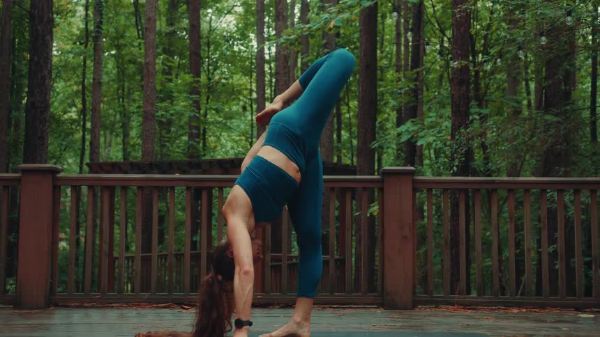 A female yogi wearing a dark blue sports bra and leggings is practicing yoga on a mat on a wooden deck in a forest transitioning through a series of balance poses including downward dog half moon and standing splits