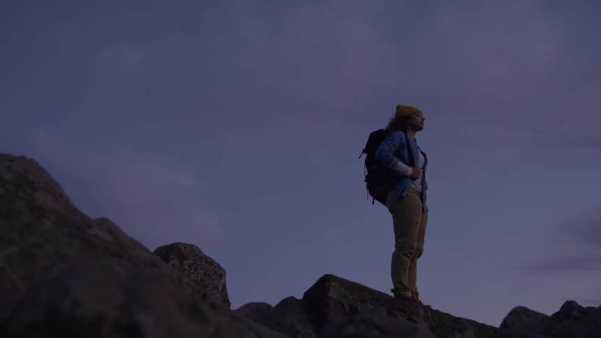 Hiker wearing a backpack stands on a mountain of rocks as she looks around